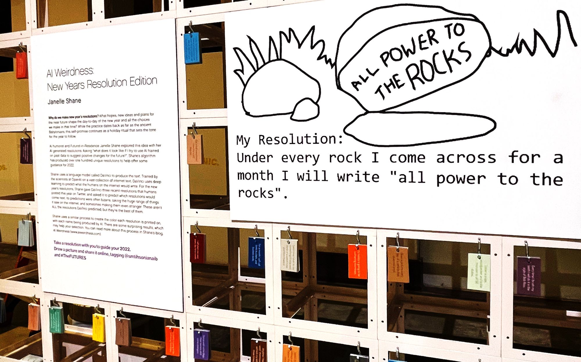Colorful tags hang from a white grid frame. Two posters are visible on the exhibit; one says "AI Weirdness New Years Resolution Edition" and the other says "My resolution: Under every rock I come across for a month I will write 'all power to the rocks'."
