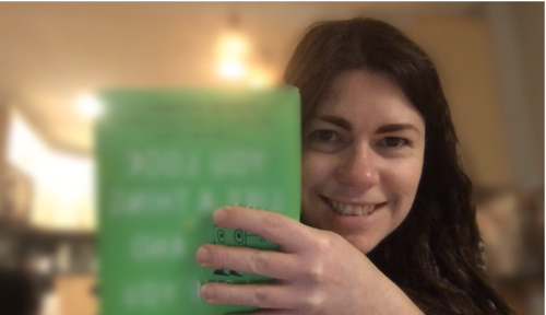 Janelle is holding up her book. The cover is blurred, except for a tiny slice between her fingers Janelle is holding up her book. The cover is blurred, except for a tiny slice between her fingers