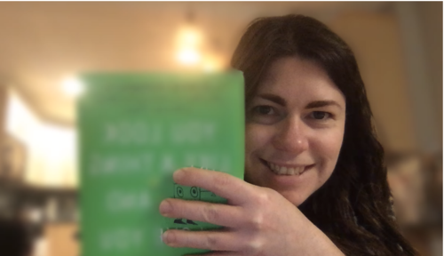 Janelle is holding up her book. The cover is blurred, except for a tiny slice between her fingers